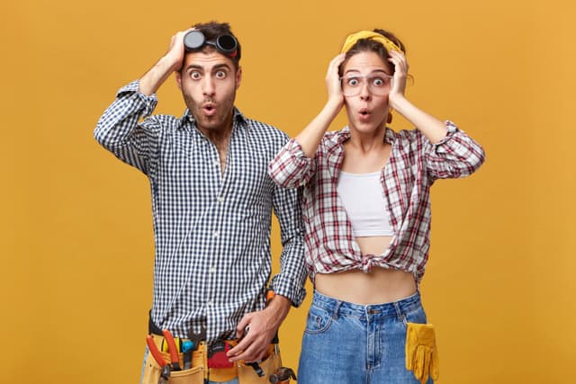 Human emotions and feelings. Two surprised astonished young Caucasian service technicians wearing safety eyeglasses and overalls having amazed and shocked looks, holding hands on their heads